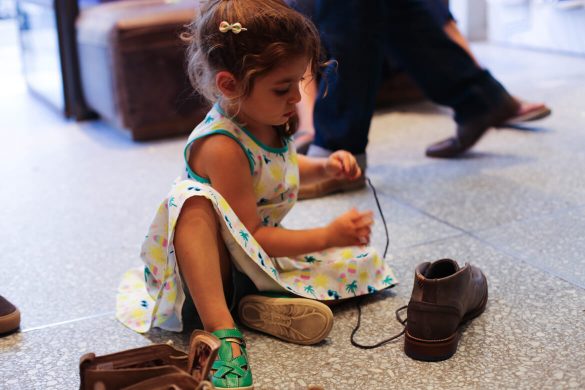 ילדה בשמלה משחקת על רצפת חנות נעליים עם שרוך נעל חומה