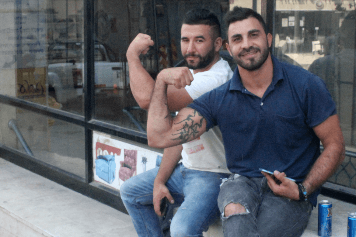 שני בחורים, אחד מהם מקועקע, אחרי שלגמו משקאות אקסל עושים שרירים לצלמת מחוץ לחנות הנעליים